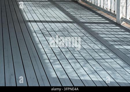 Sunlight and shadows from railings create a geometric pattern on a clean wooden terrace floor, evoking a peaceful and inviting atmosphere. Stock Photo