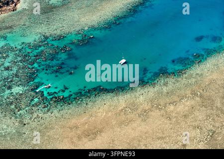 Aerial view of a white sailing yacht moored in deep turquoise water beside a shallow reef flat and coral heads, off the Australian coast Stock Photo