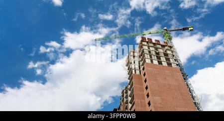 High rise building under construction rises beside tower crane against dramatic blue sky, leaving wide copy space for banner. Stock Photo