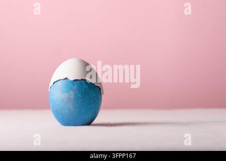 Blue Easter egg wears cracked white shell cap, minimalist pastel still life with gentle humor and copy space on pink background. Stock Photo