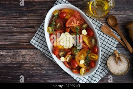 Colorful tomatoes arranged with mozzarella fresh basil olives and garlic in a white serving dish on a rustic wooden table Stock Photo