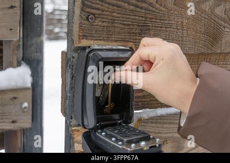 Hand places keys inside outdoor keypad key lockbox on wooden fence in winter snow to store spare access for rental guests. Stock Photo