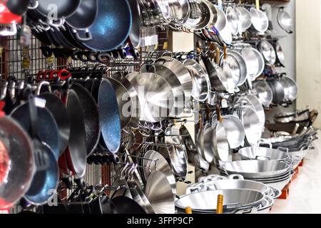 A diverse collection of frying pans and woks displayed on metal racks in a kitchenware store. The variety of materials and sizes is evident. Stock Photo
