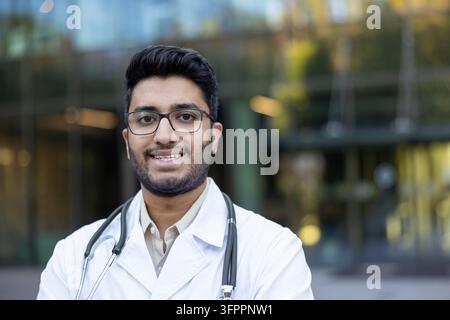 Young indian male doctor wearing a white lab coat and stethoscope, smiling confidently while standing outdoors, representing healthcare, medicine, and medical professionals Stock Photo