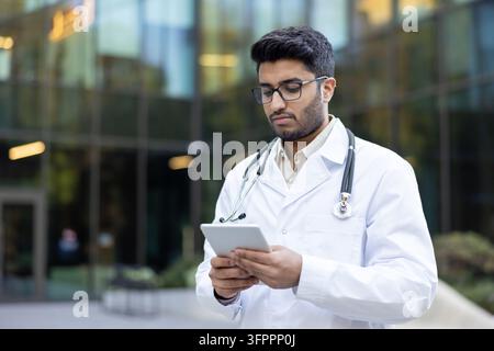 Young male doctor wearing a white lab coat and stethoscope, standing outdoors while focused on a digital tablet, symbolizing modern medical technology and data management in healthcare Stock Photo