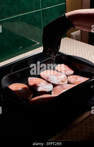 Close up of gloved hand sprinkling coarse salt over fresh tuna steaks arranged in black tray on kitchen counter, preparing raw fish for cooking, salt Stock Photo