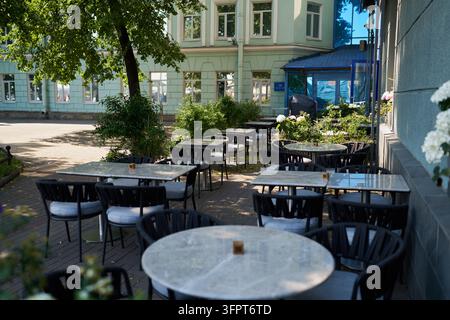 Sunny outdoor cafe terrace with empty round tables and woven chairs under large leafy tree beside mint green building facade with hydrangea blooms Stock Photo