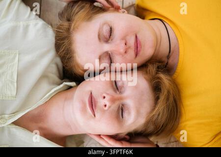 Two red-haired sisters lie cheek to cheek with their eyes closed. Similar women with fair skin and freckles. Beautiful twin women. Stock Photo