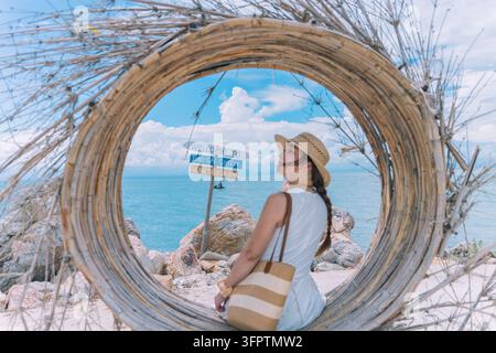 Woman in straw hat with striped tote bag sitting in bamboo ring beside Welcome to Dao Cai Chien sign, Cai Chien Island, Quang Ninh, Vietnam Stock Photo