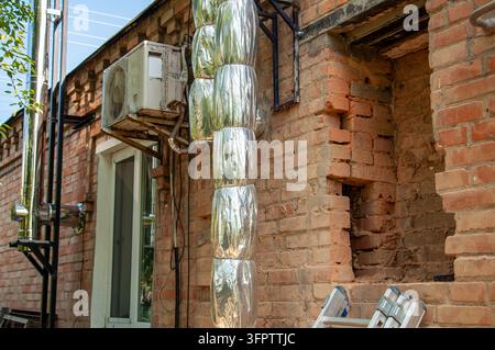 An outdoor air conditioning unit is mounted on a brick wall next to insulated pipes and a window Stock Photo