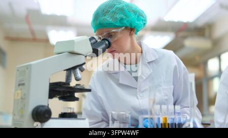Female scientist wearing a lab coat and safety goggles observing samples with a microscope, performing biological research and development in a modern Stock Photo