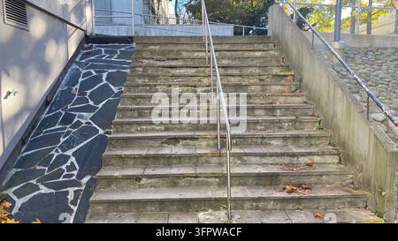 Weathered outdoor concrete staircase with metal handrail and narrow stone paved ramp beside building Stock Photo