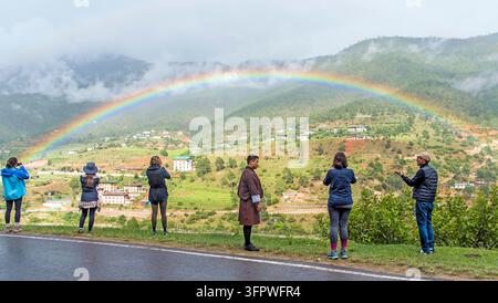 Bhutanese man in traditional gho robe with tourists photographing a vibrant rainbow arching over lush green Punakha Valley during the monsoon. Stock Photo