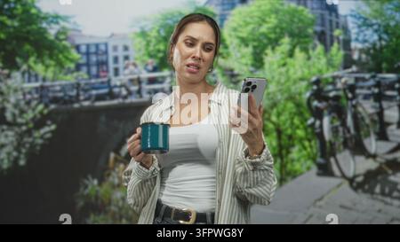 Woman holds teal mug and smartphone, frowning while checking messages, wearing white tank top and striped overshirt near canal with bicycles on a stre Stock Photo