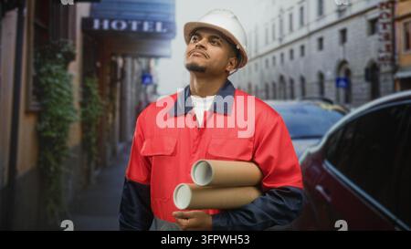 Hispanic man in hard hat holding rolled blueprints and wearing red uniform, beard visible, looks up on street near hotel; determination planning. Stock Photo