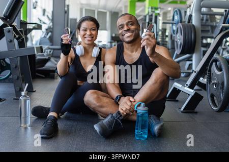 Smiling black couple sitting on gym floor showing protein bars to camera Stock Photo