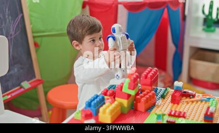 Boy holding white and blue toy robot with both hands at a playroom table strewn with colorful building blocks and a small tent; curiosity imagination. Stock Photo