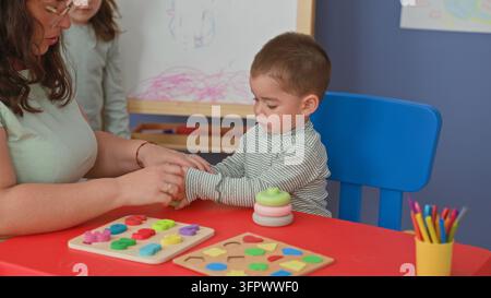 Woman helps toddler stack colorful rings and grip toy on red table in preschool building; concentration learning. Stock Photo