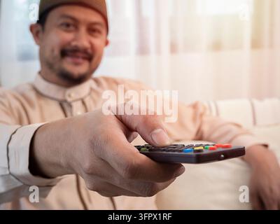 Enjoy television program. Happy young Asian muslim man watching TV switching channels with remote control sitting on sofa at home. Stock Photo
