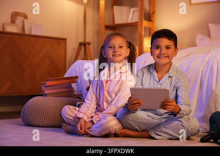 Digital childhood and remote learning, happy children in pajamas using a tablet computer for entertainment or education in a cozy bedroom at night Stock Photo