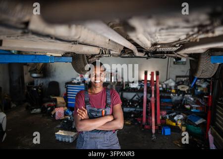 Confident African Female Mechanic Smiling Under Lifted Car in Garage Stock Photo