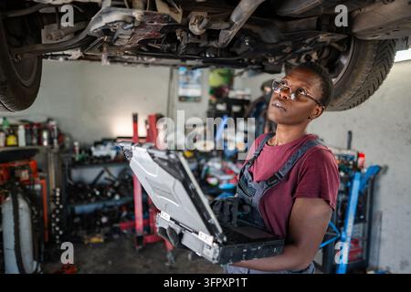 Young African Female Mechanic Inspecting Car Using Laptop in Garage Stock Photo
