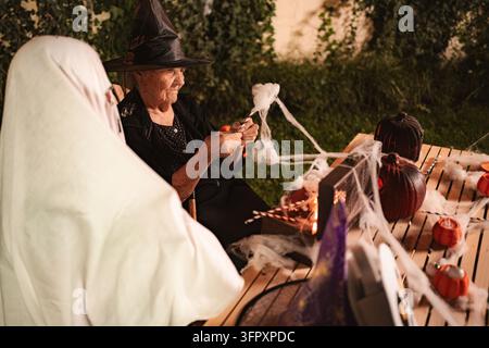 Elderly woman making Halloween decorations with her friend dressed as a ghost Stock Photo