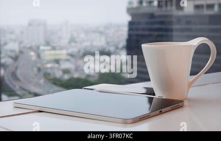 Close up of a modern workspace setup featuring a digital tablet, mobile phone, and a white coffee mug on a reflective table. The background shows a bl Stock Photo