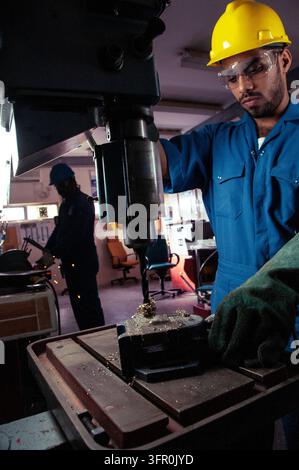 Skilled factory worker wearing protective gear operating a vertical drilling machine while a colleague works in the background area Stock Photo