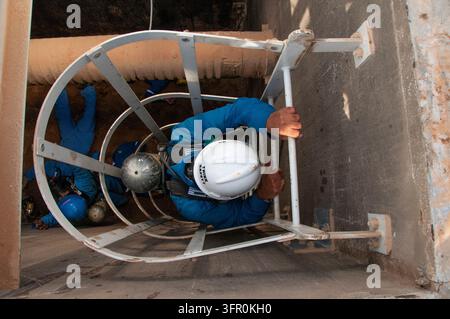 Workers on a rescue mission wearing safety gear, including a hard helmet written Think Safety and breathing apparatus, climbing down a caged ladder Stock Photo