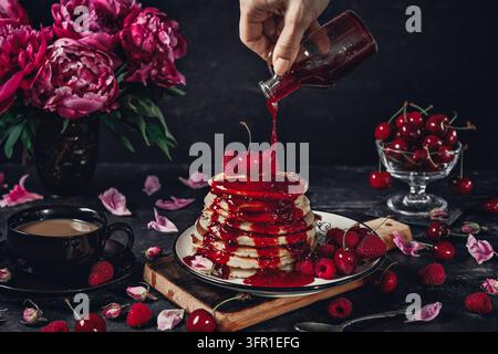 Stack of pancakes with cherry sauce being poured over them, fresh cherries and pink peonies on a dark rustic table Stock Photo