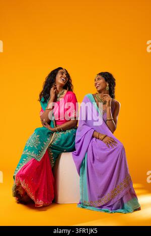 Diverse female friends sitting on white stool against orange backdrop wearing saris and jewelry Stock Photo