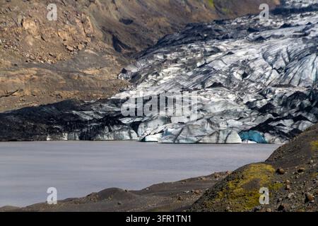Close view of a melting glacier meeting a calm gray lake beneath steep rocky cliffs, showing dark ice, cracks, and small blue ice caves. Stock Photo