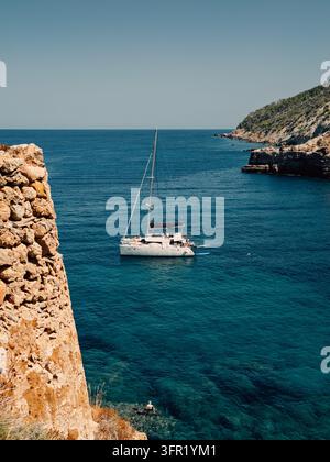 A sailboat is captured sailing on the vibrant turquoise waters of the Mediterranean Sea, framed by rugged, rocky coastal cliffs. The clear blue sky in Stock Photo