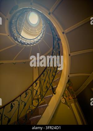 A low-angle view of an elegant spiral staircase within a historic building, leading upwards towards a circular skylight. The ornate wrought iron raili Stock Photo