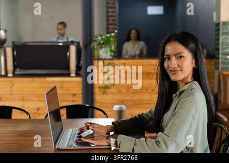 Mid-adult Asian woman typing on laptop at cafe table, wearing light olive shirt, coffee cup nearby Stock Photo