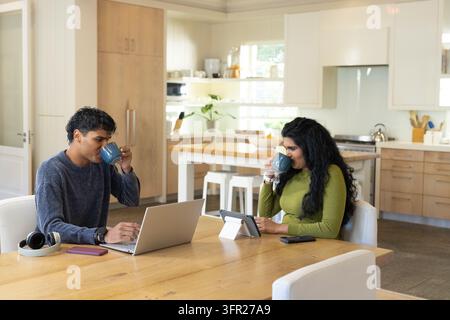 Diverse couple sitting at wooden dining table in modern kitchen, using laptop, tablet and blue mugs Stock Photo