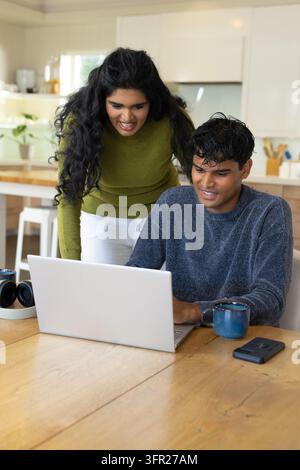 Diverse couple leaning over laptop while man typing at wooden table with blue mug and smartphone Stock Photo