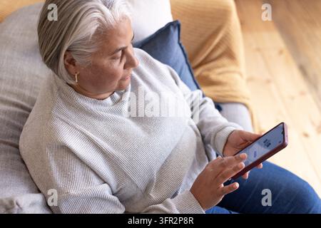 Senior African-American woman sitting in gray sweater on couch at home holding purple smartphone Stock Photo