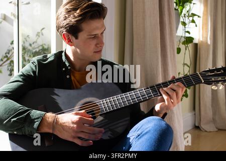 Man sitting on floor by large window wearing dark shirt and jeans, playing black acoustic guitar Stock Photo