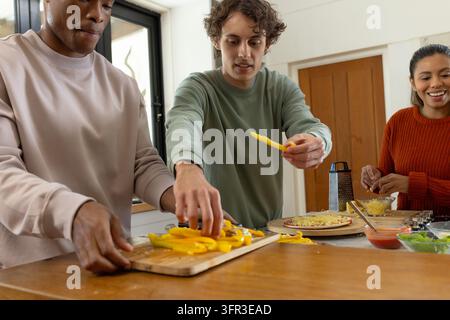 Diverse friends chopping yellow peppers on board and topping pizzas on peel at kitchen island Stock Photo