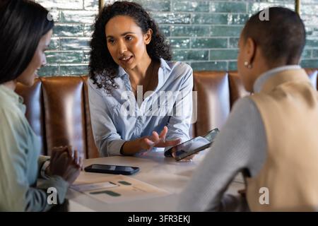 Diverse women coworkers discussing project at cafe booth in business wear holding tablet and charts Stock Photo