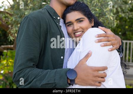 Diverse couple embracing on porch by foliage, woman in white blouse, man wearing wristwatch Stock Photo