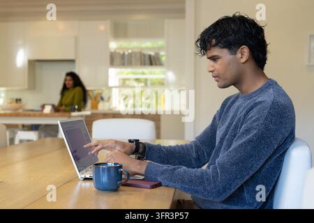 Diverse couple showing man wearing blue sweater working on laptop at table with mug and phone Stock Photo