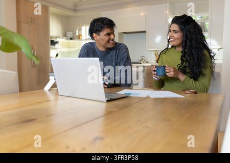 Diverse couple sitting at wooden dining table, holding mugs, discussing documents near laptop Stock Photo