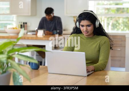 Diverse couple in kitchen, woman working on silver laptop in green sweater and headset, copy space Stock Photo