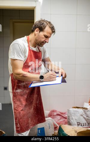 Male butcher in a stained red apron writes on a clipboard beside packaged meat in a tiled shop, checking stock and preparing an order. Stock Photo