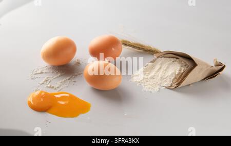 Brown Eggs with Spilled Yolk, Flour and Wheat Ear on White Surface Stock Photo