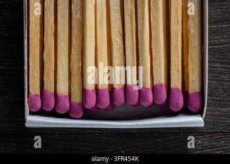 Matches with pink heads in cardboard box close up. Top view of wooden matches with purple sulfur heads inside a matchbox. Row of matchsticks on dark b Stock Photo