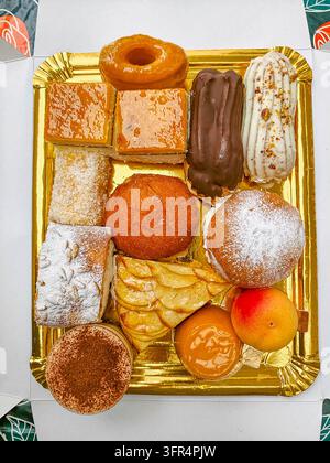 Flat lay top-down photograph of a gold rectangular tray packed with diverse desserts, cakes, tiramisu, and fruit tarts on a white box background. Rich Stock Photo